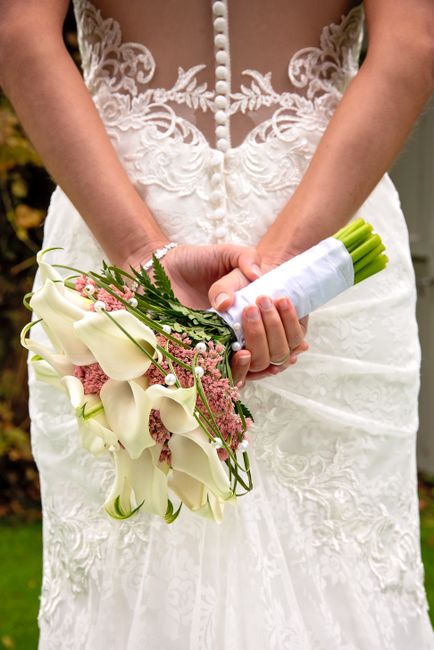 A bride in a white lace wedding gown is holding a bouquet of fresh white calla lilies and pink, green, and white accent flowers, wrapped in white ribbon. She stands outdoors, with the floral arrangeme