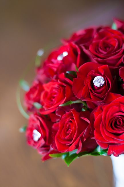 A close-up of a bouquet of fresh red roses with tightly layered petals, accented by sparkling rhinestone embellishments nestled among the blooms. The bouquet is wrapped in elegant white paper and is h