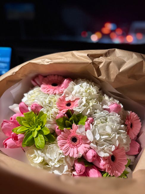 A bouquet of fresh flowers comprising white hydrangeas, pink gerbera daisies, and green foliage, wrapped in brown craft paper and white tissue paper, positioned in a delivery setting. The flowers exhi