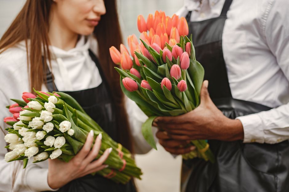 A woman and a man, both dressed in white shirts and black aprons, are engaged in a flower delivery process at a florist shop. The woman is holding a bouquet of white tulips with fresh green stems and 